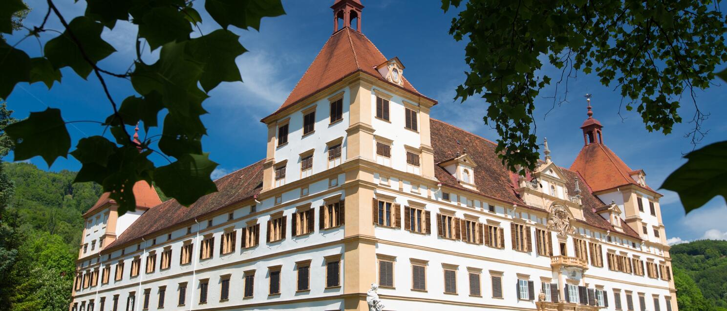 Schloss Eggenberg in Graz, weißgelbe Fassade mit rotem Dach, Statuen vor dem Eingang, bewaldete Hügel dahinter.