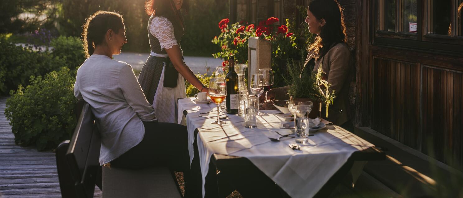 Waitress in dirndl serving two guests at a set table on a wooden terrace in evening sunlight.