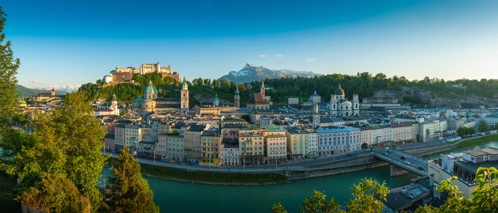 Panoramic view of Salzburg's old town with Hohensalzburg Fortress, cathedral, Salzach River and snow-capped peak in the background.