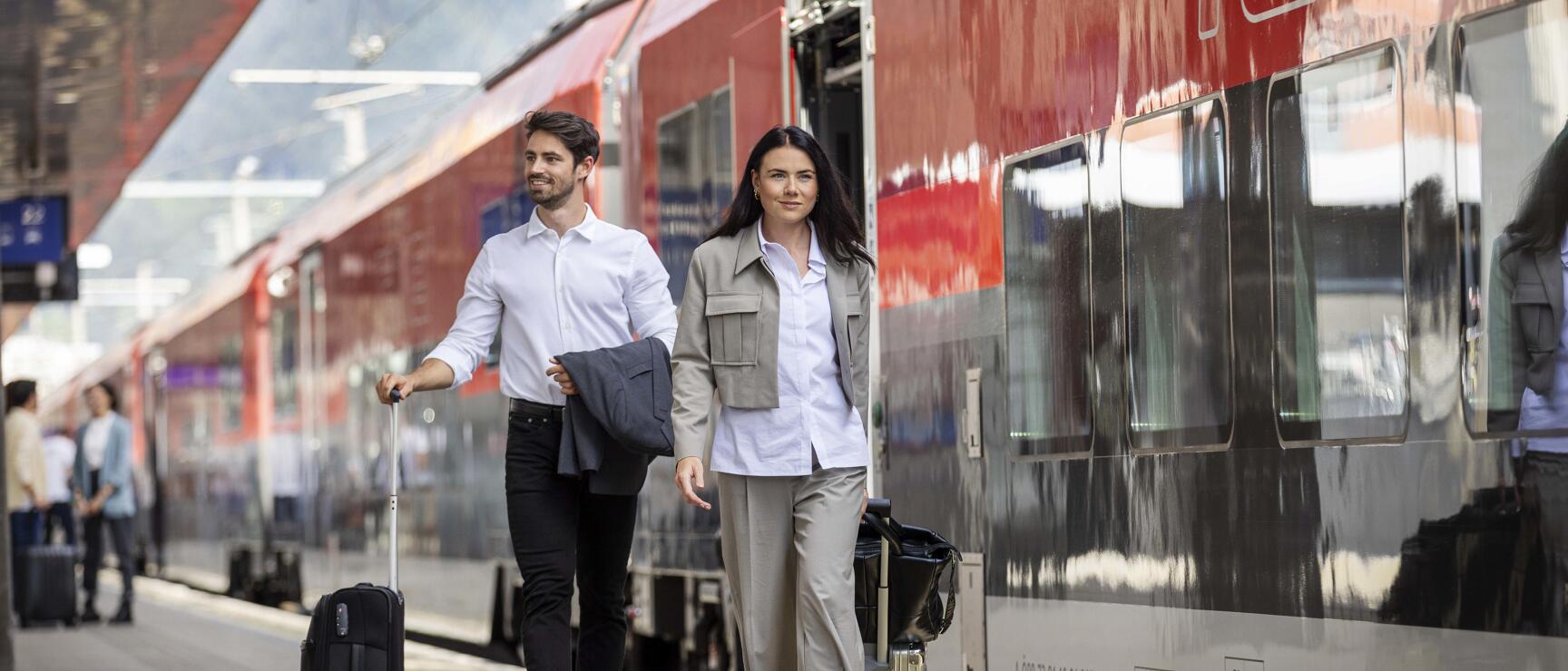 Two people with trolleys walking along red ÖBB train on railway platform.