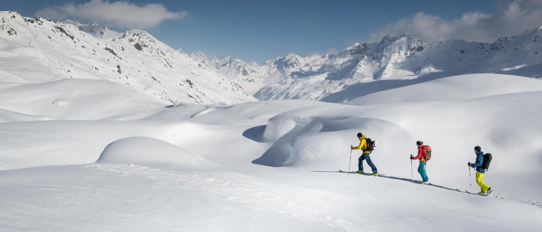 Three ski tourers with backpacks in snow-covered high mountain landscape, snow-capped peaks in background.