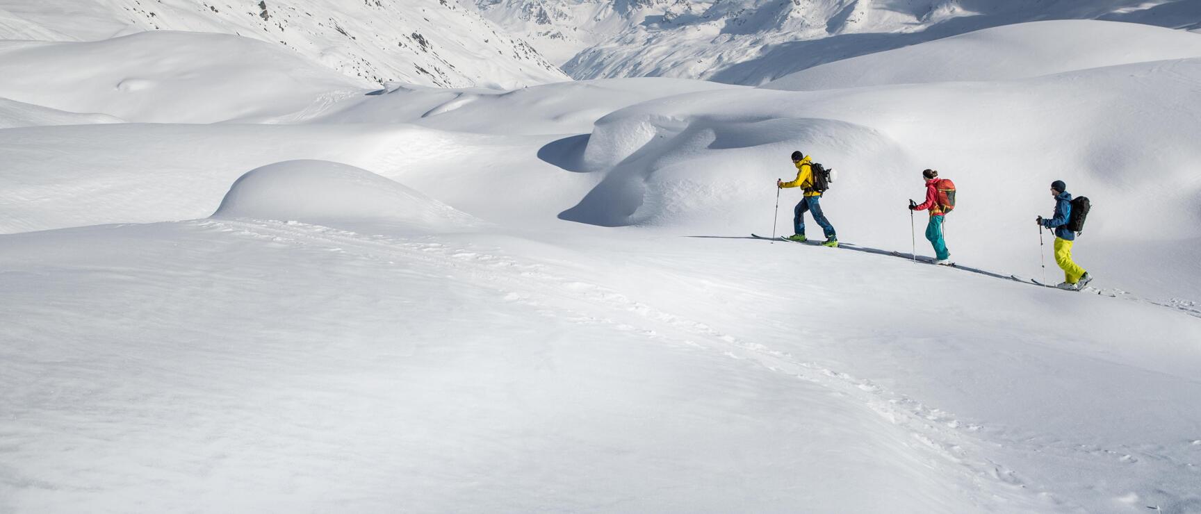 Drei Skitourengeher mit Rucksäcken in verschneiter Hochgebirgslandschaft, schneebedeckte Gipfel im Hintergrund.