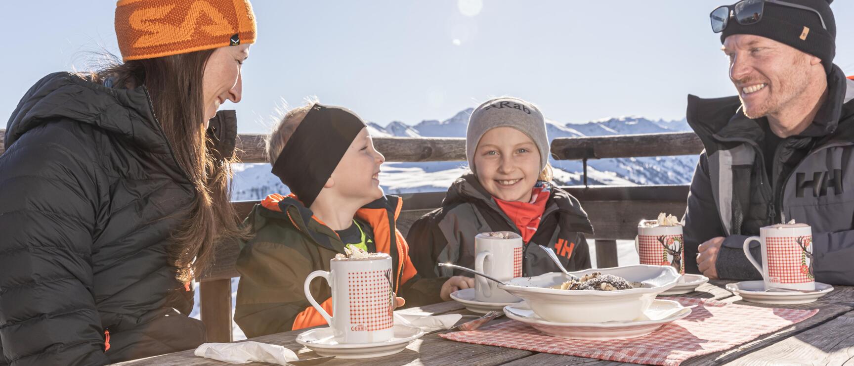Two adults and two children at a wooden table in a mountain hut, hot drinks and Kaiserschmarrn, snow-covered mountains.