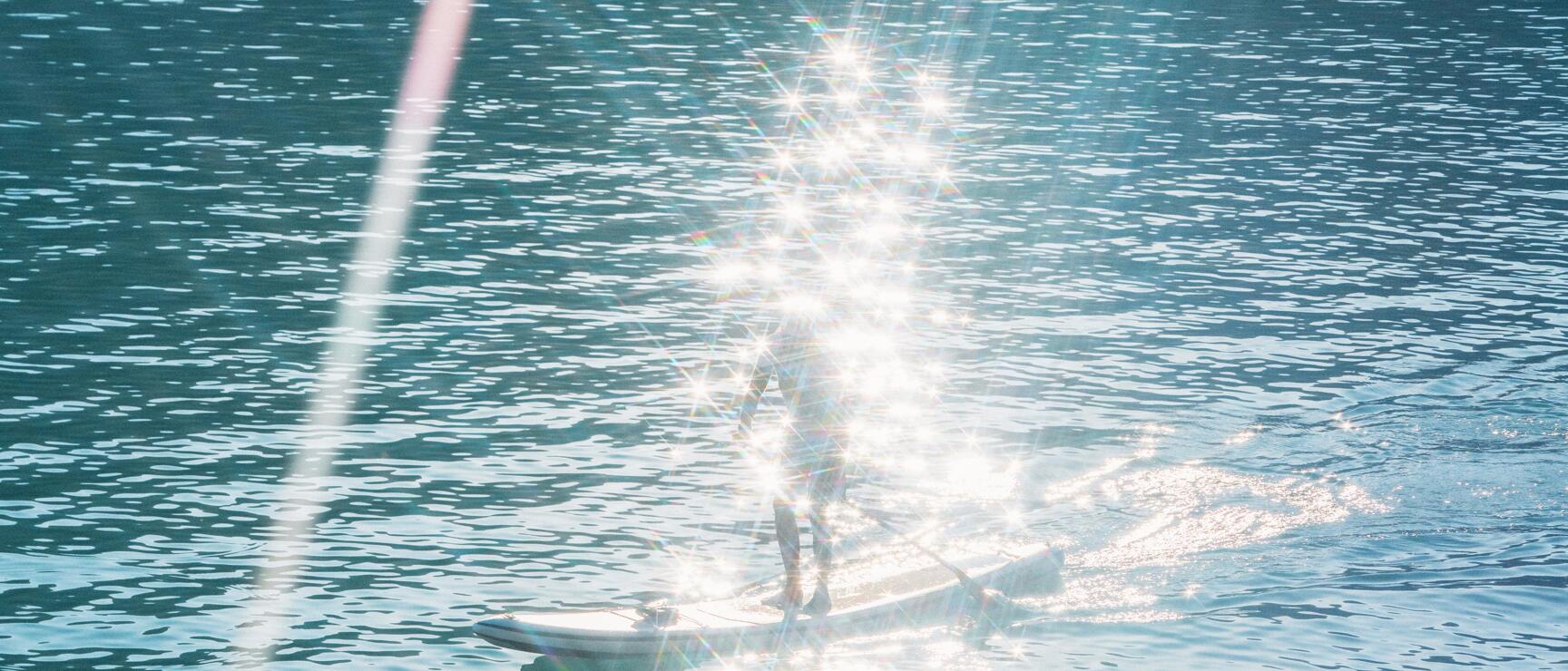 Person on stand-up paddleboard on turquoise blue lake with strong sunlight reflections and green shore in foreground.