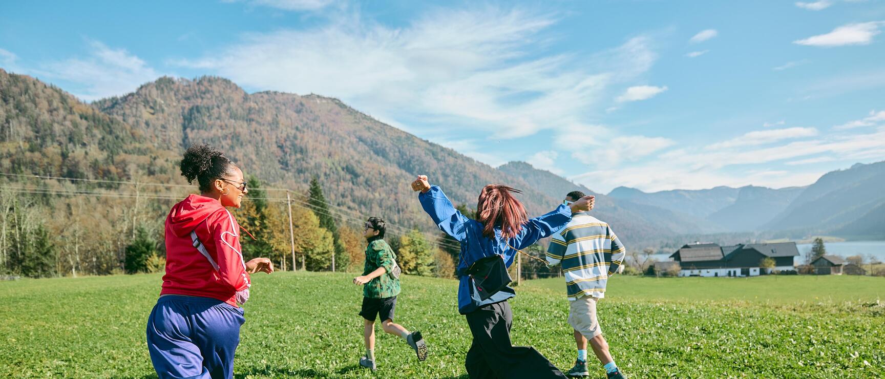 Vier Personen laufen über eine grüne Wiese, Bergpanorama und See im Hintergrund, sonniger Herbsttag.
