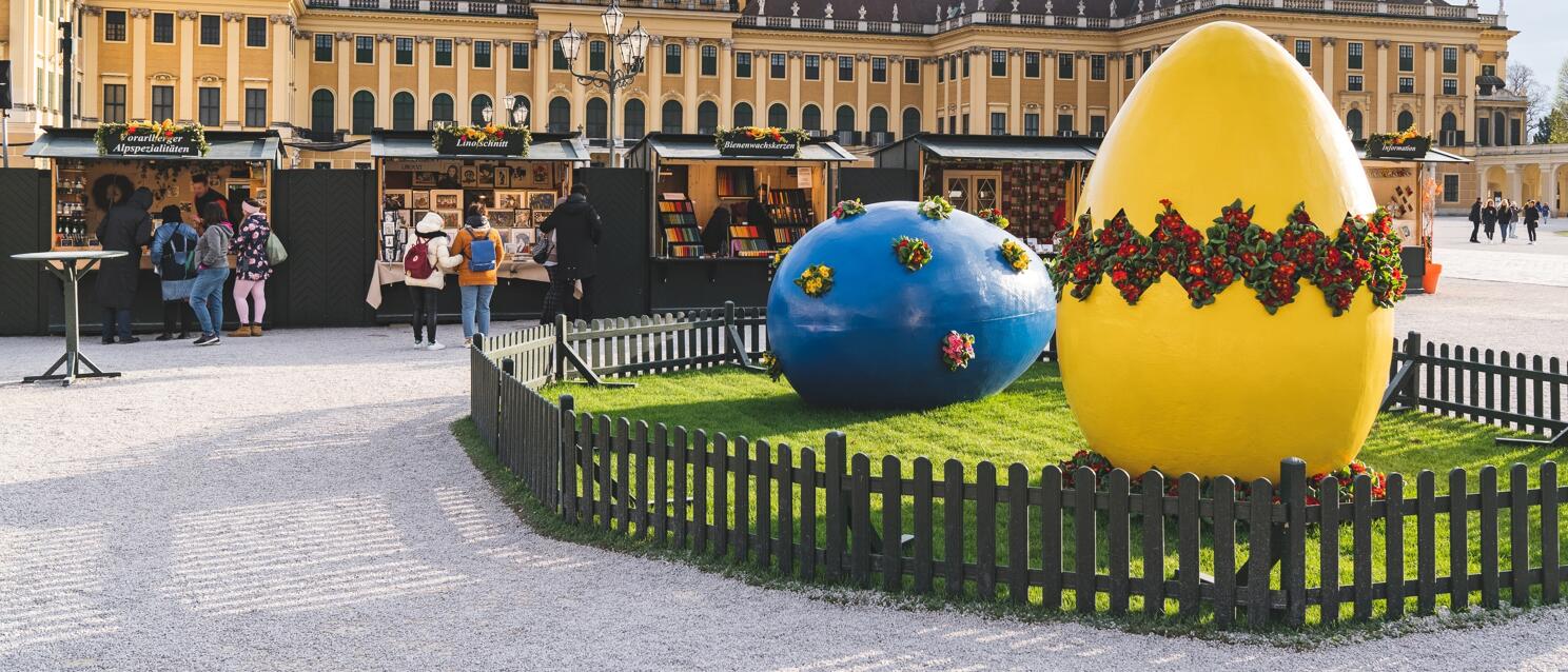 Easter market in front of Schönbrunn Palace, Vienna: two large giant eggs in blue and yellow with flower decorations, market stall in background.