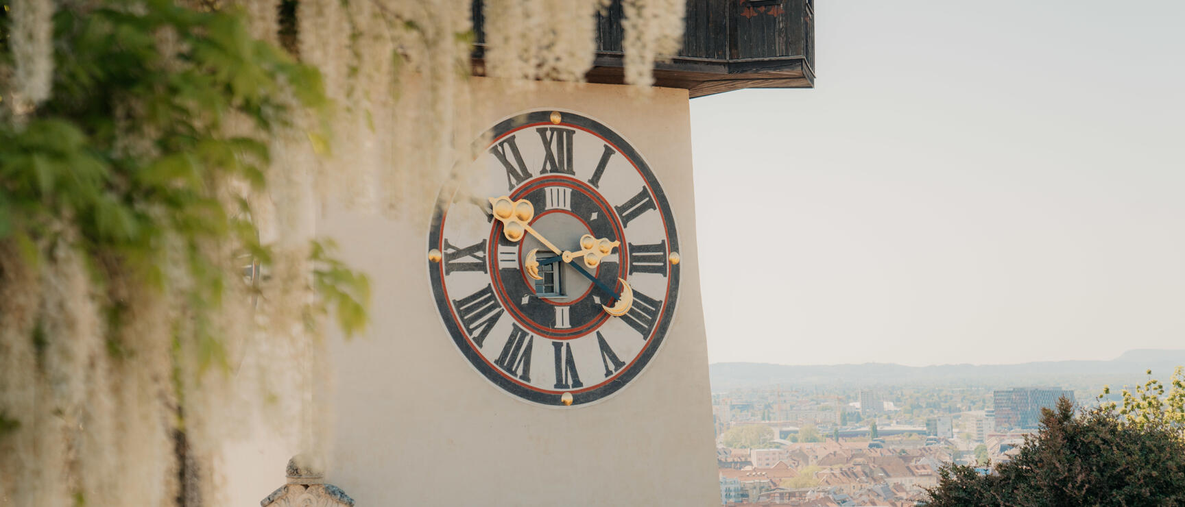 Clock tower on Schlossberg in Graz with large clock face, city view in the background.