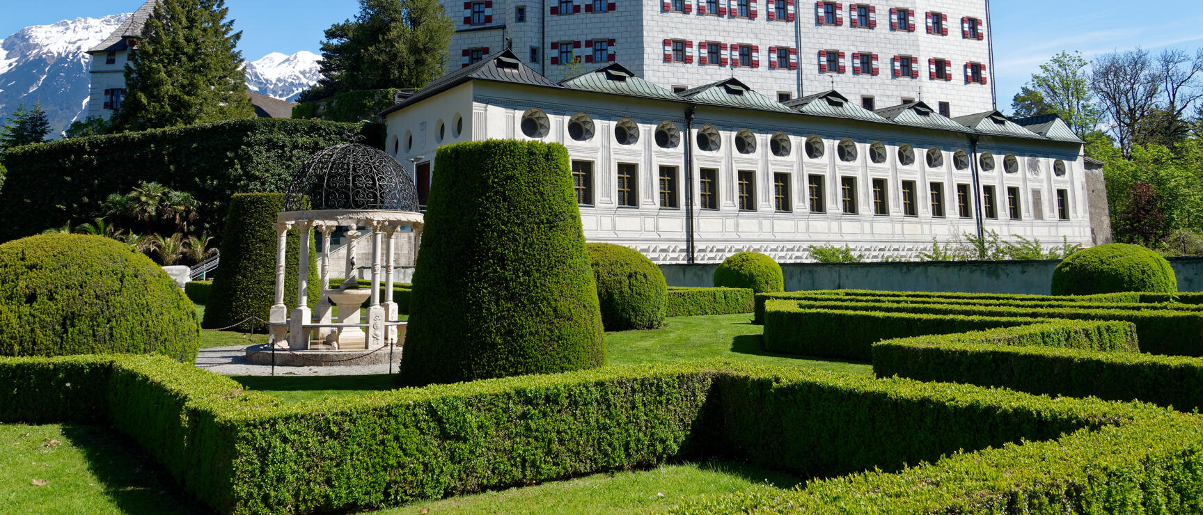 Ambras Castle in Innsbruck with geometrically cut garden, pavilion and snow-covered mountains in the background.