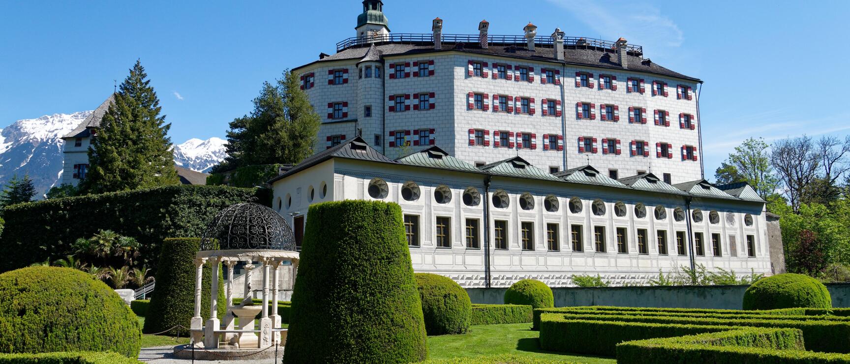 Ambras Castle in Innsbruck with geometrically cut garden, pavilion and snow-covered mountains in the background.