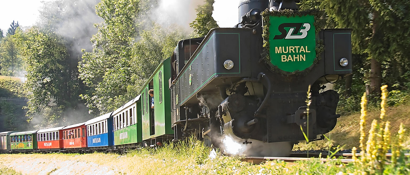 Steam locomotive of the Murtal Railway with colourful carriages driving through summer forest landscape, smoke from chimney.