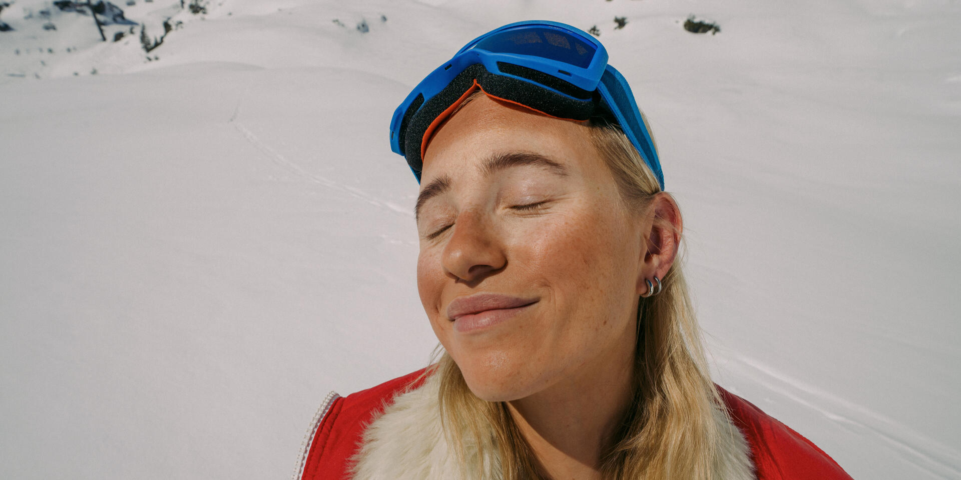 Woman with blue ski goggles on head and red jacket, eyes closed, snow-covered mountain in background.
