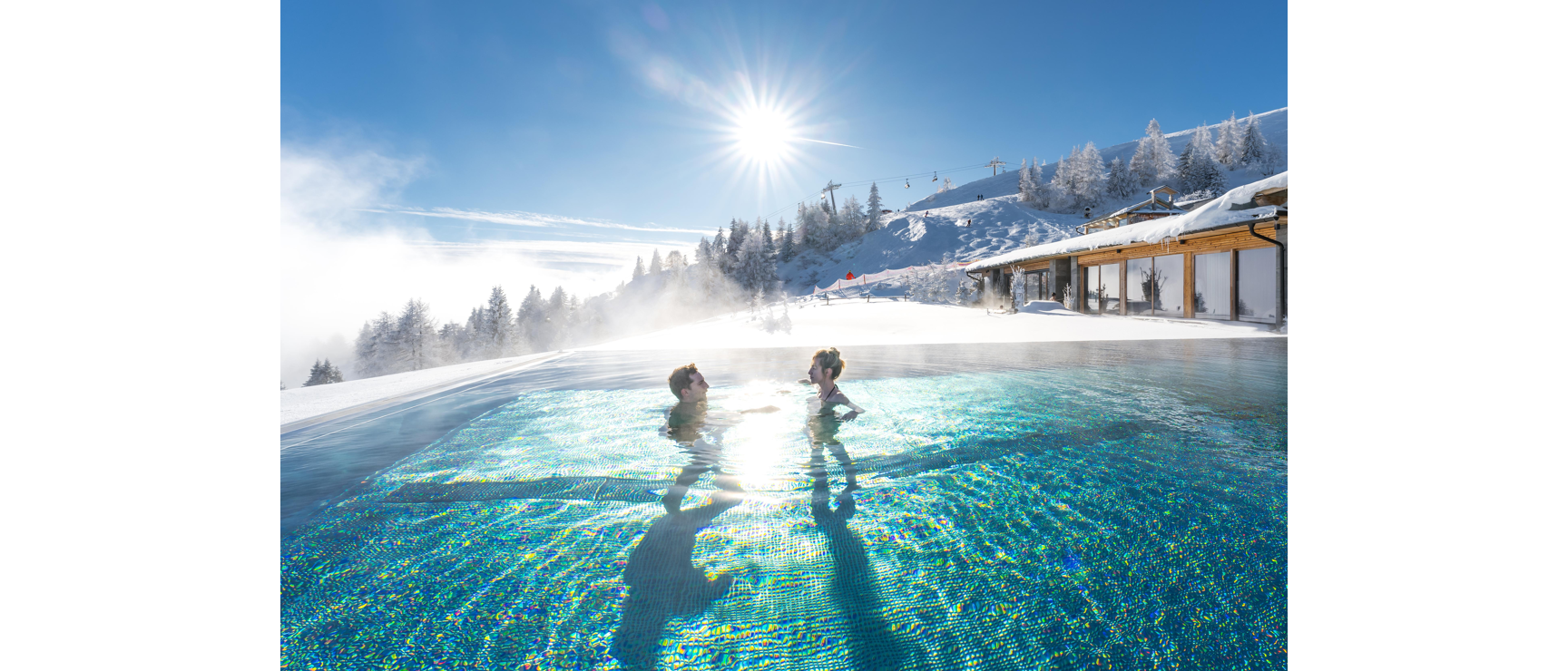 Two people in a steaming outdoor pool, snowy ski slope with chairlift and wooden building in background.