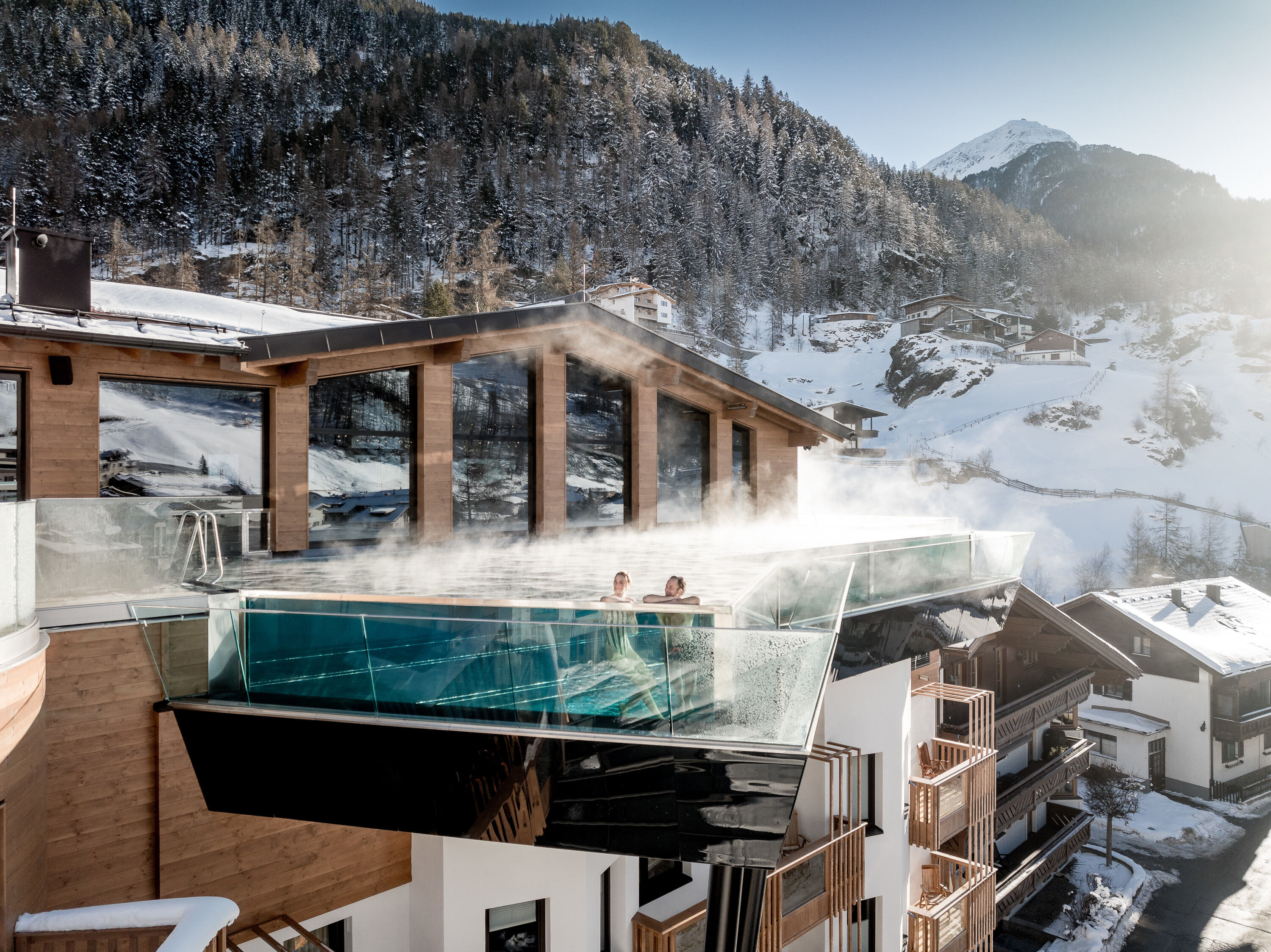 Two people in steaming infinity pool of hotel building, snow-covered mountain landscape in background.