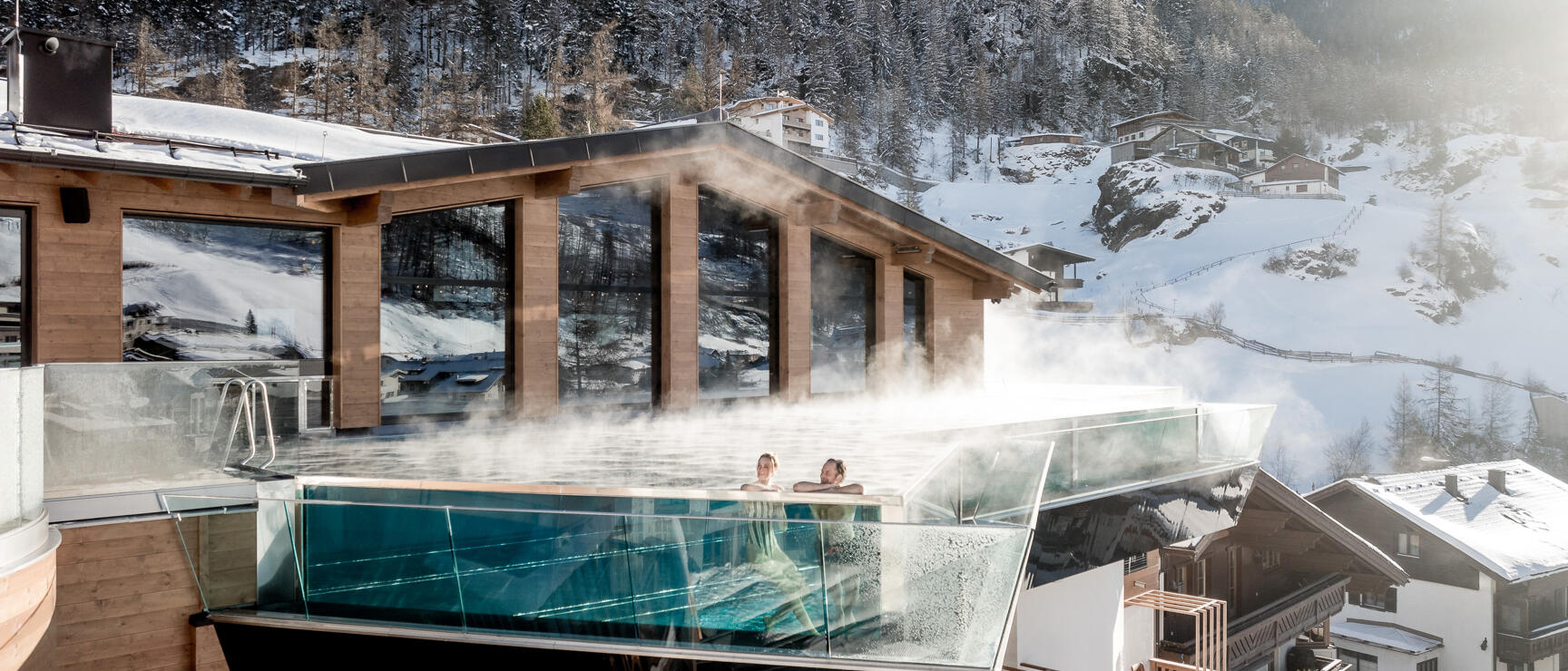Two people in steaming infinity pool of hotel building, snow-covered mountain landscape in background.