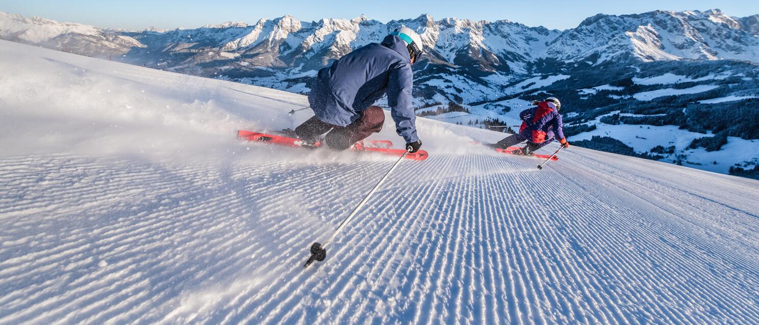 Two skiers on a groomed slope with snow-covered mountain range and valley in the background, blue sky.