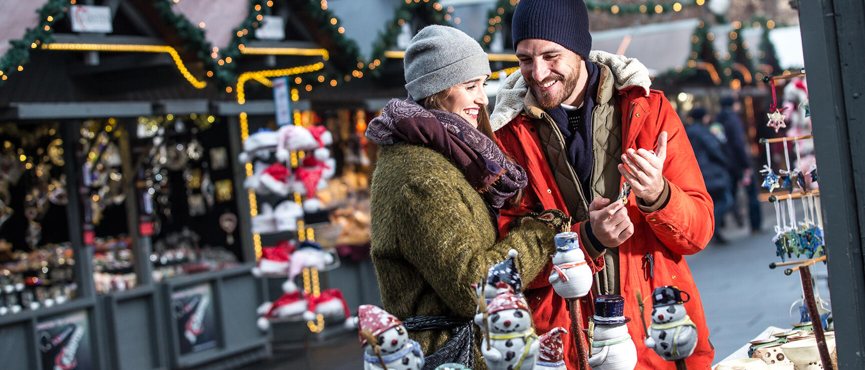 Couple at a Christmas market stall with snowman ceramic figurines, illuminated huts in the background.