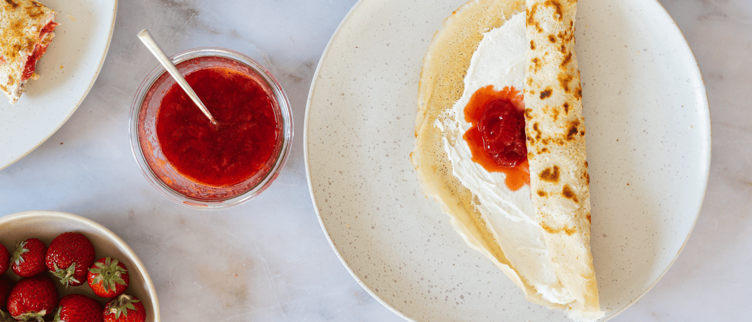 Pancake with whipped cream and strawberry jam on plate, beside glass of jam and bowl of strawberries.