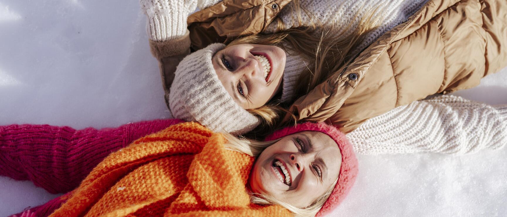 Two laughing women in winter hats and scarves lying in the snow, bird's eye view.