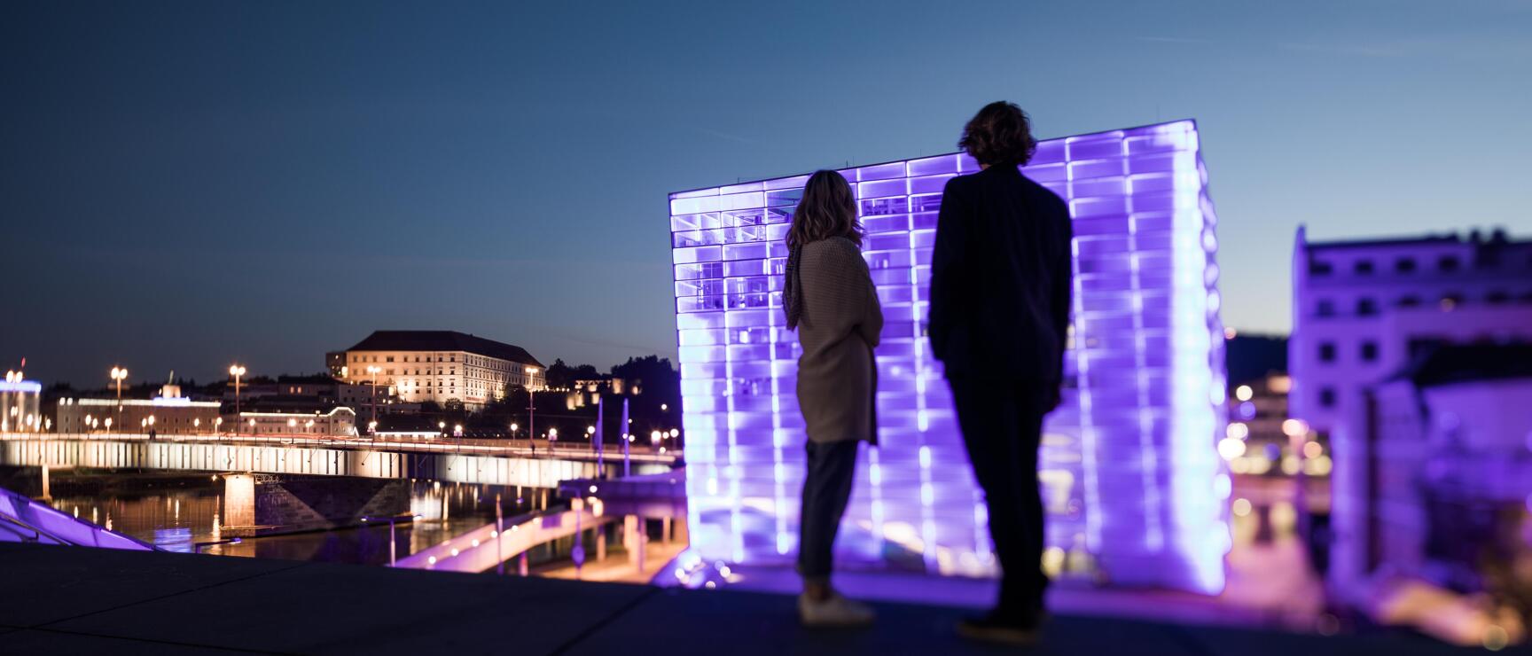 Two people in front of purple-lit glass cube at night, Danube riverbank and Linz bridge in background.