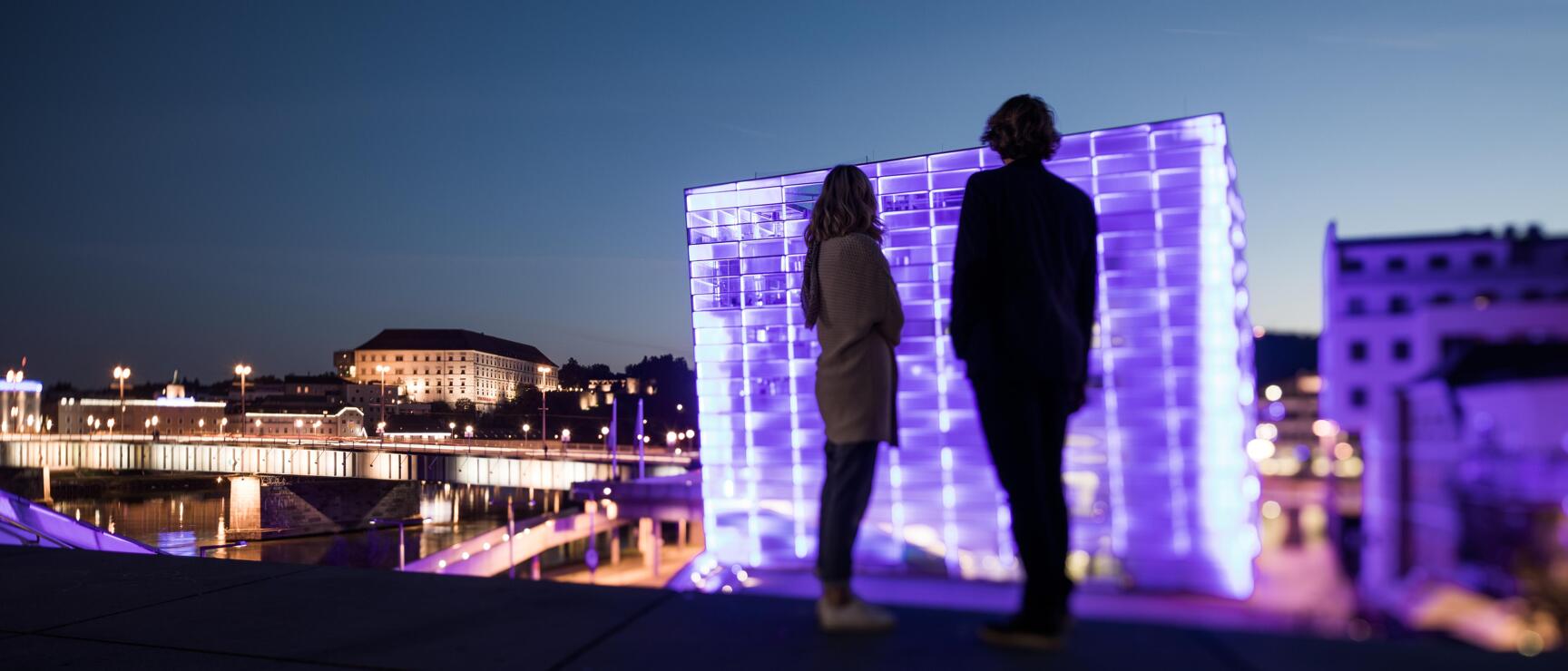 Two people in front of purple-lit glass cube at night, Danube riverbank and Linz bridge in background.