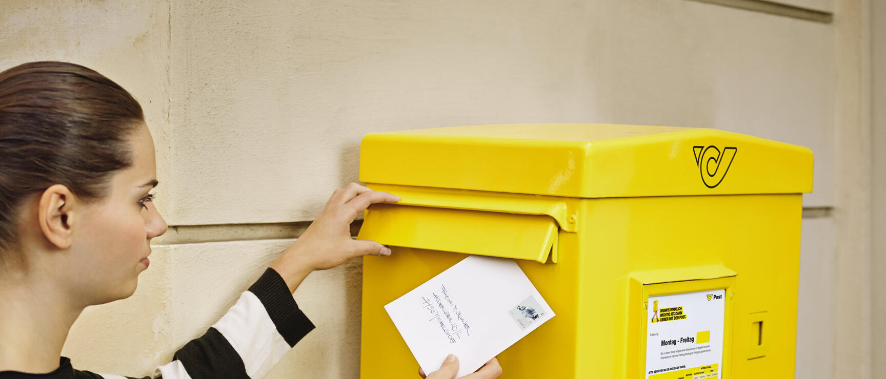 Woman posting a letter into a yellow Austrian mailbox on a house wall.