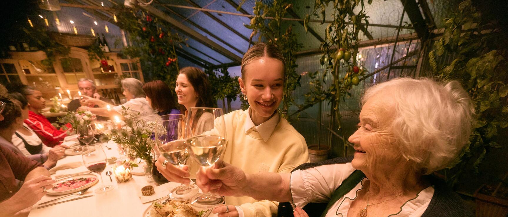 Two women toasting with white wine at a set table in a green plant-filled greenhouse restaurant.