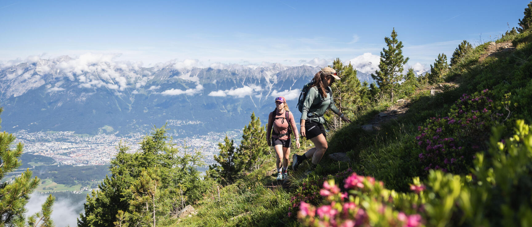 Two female hikers on a mountain path above Innsbruck, alpine roses in the foreground, mountain panorama in the background.