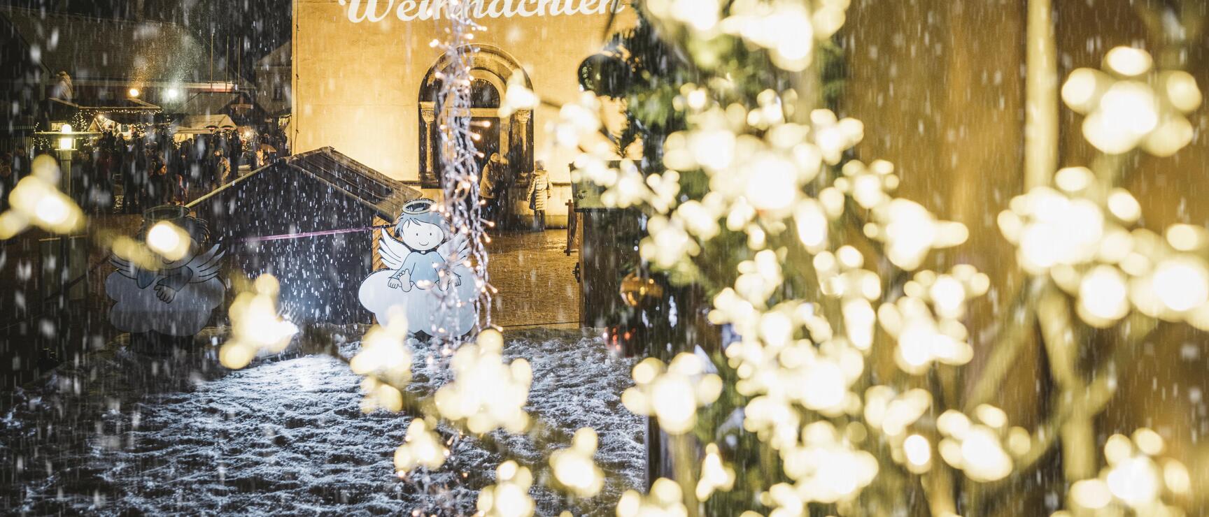Christmas market during snowfall with 'Merry Christmas' sign on church wall and festive lights in foreground.