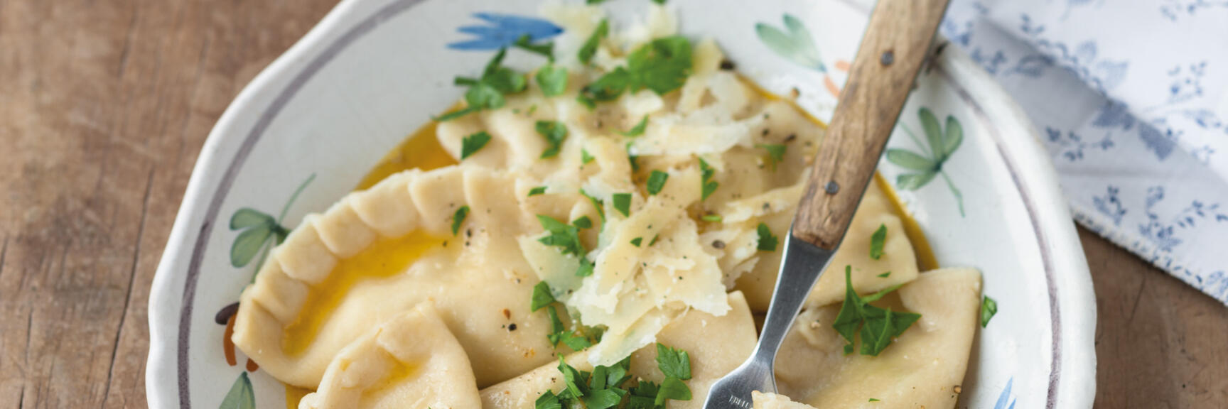 Kärnter Kasnudeln mit Butter, Käse und Petersilie in gemusterter Schüssel, Gabel, Holztisch.