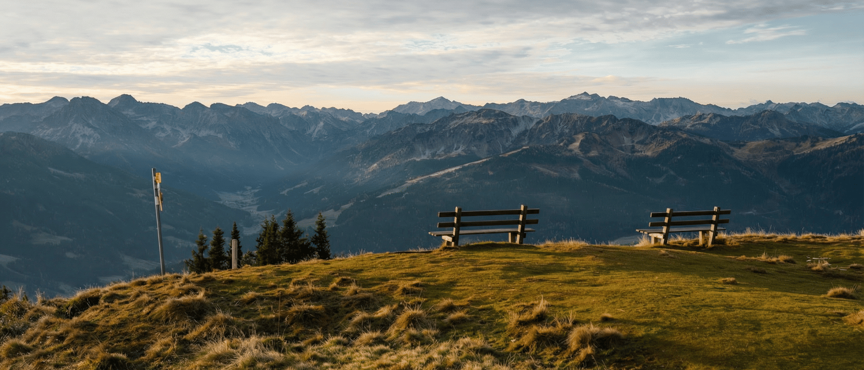 Two wooden benches on an alpine meadow with panoramic mountain range view, overcast sky.