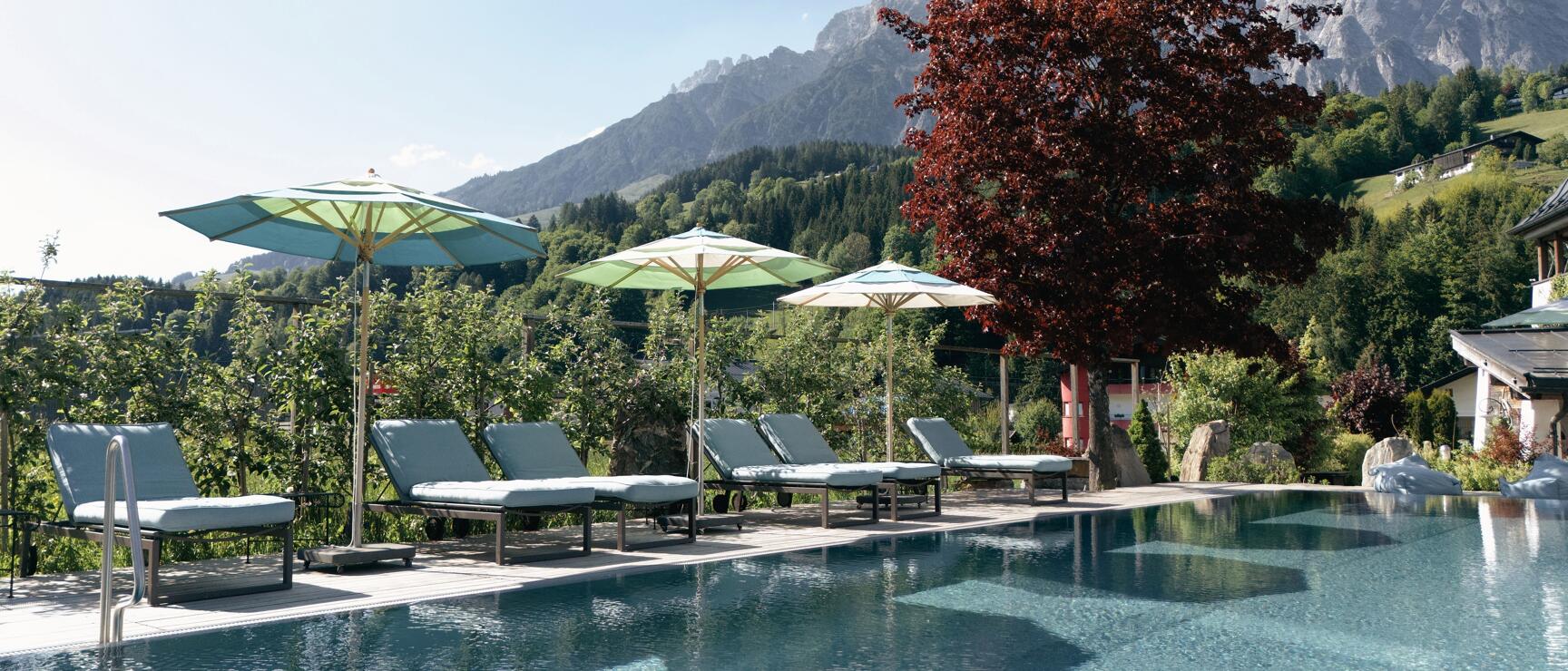 Hotel pool with loungers and green umbrellas, rocky peak and red tree in background.