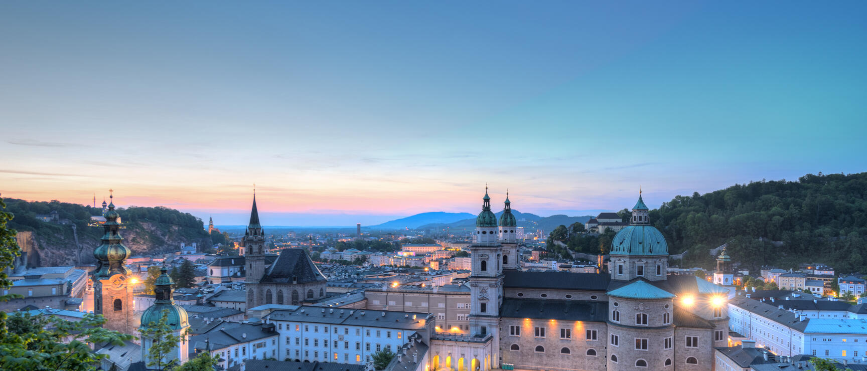 Illuminated Salzburg Old Town at evening light with cathedral and church towers, hills in background.