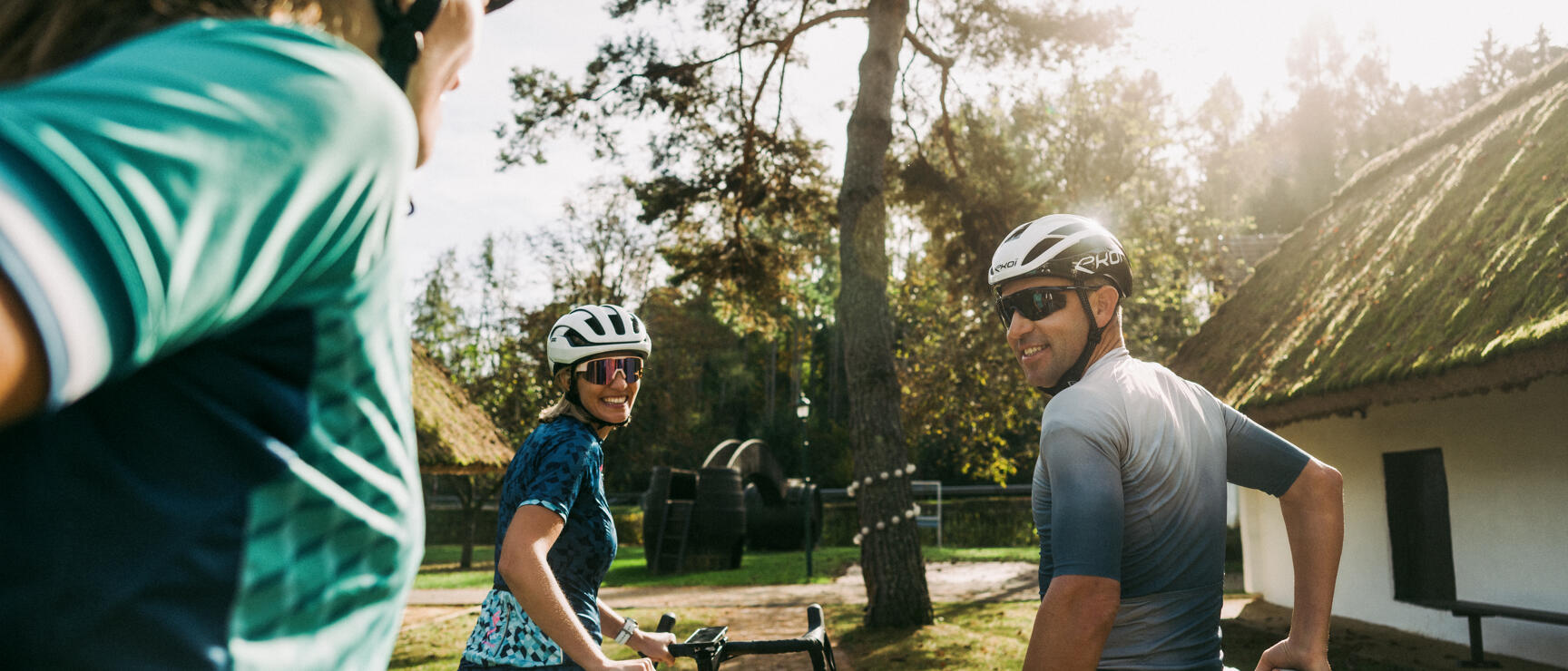 Drei Radfahrer mit Helmen und Sonnenbrillen machen Pause bei einem Reetdachhaus im Sonnenlicht.