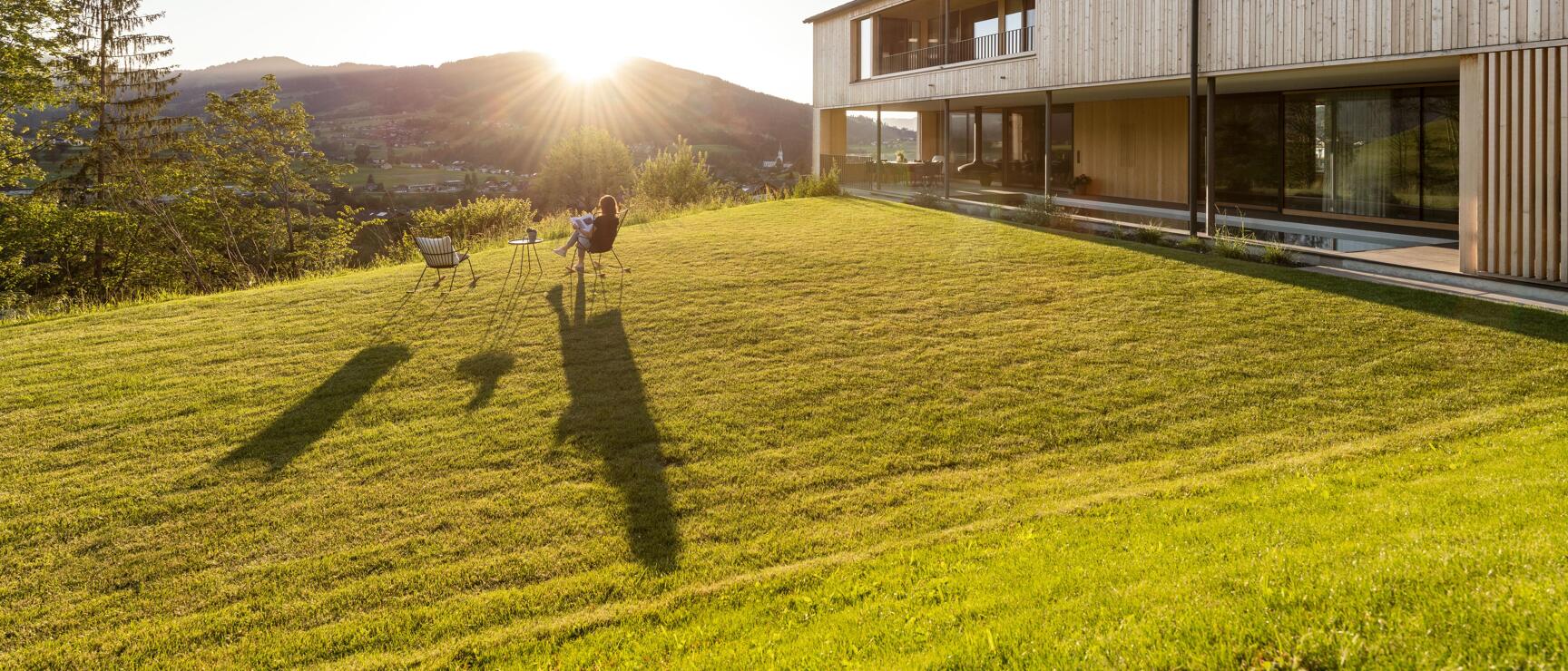 Person sitting on chair in front of modern wooden building on green meadow, sunset behind hills in background.