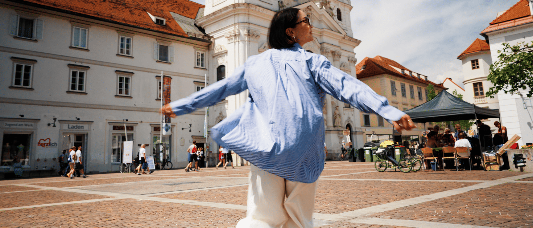 Frau mit ausgebreiteten Armen auf einem Stadtplatz vor einem barocken Kirchturm, Graz.
