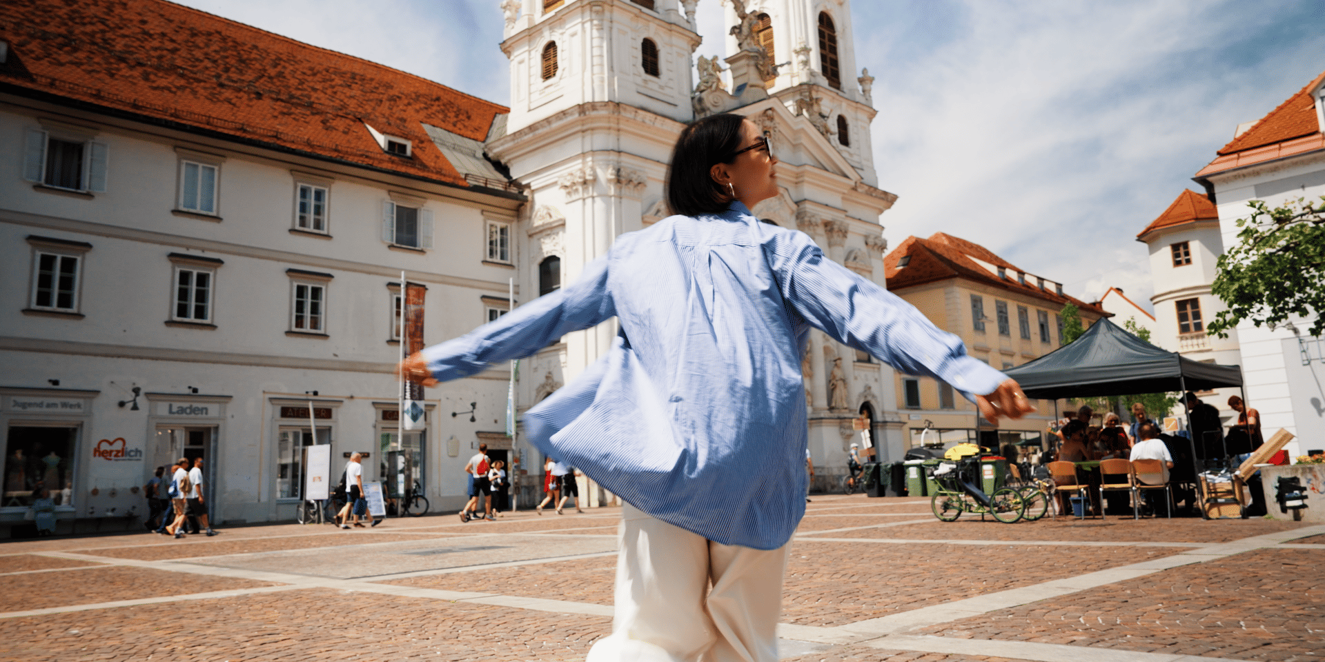 Woman with outstretched arms on a city square in front of a baroque church tower, Graz.