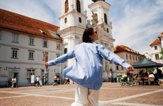 Woman with outstretched arms on a city square in front of a baroque church tower, Graz.