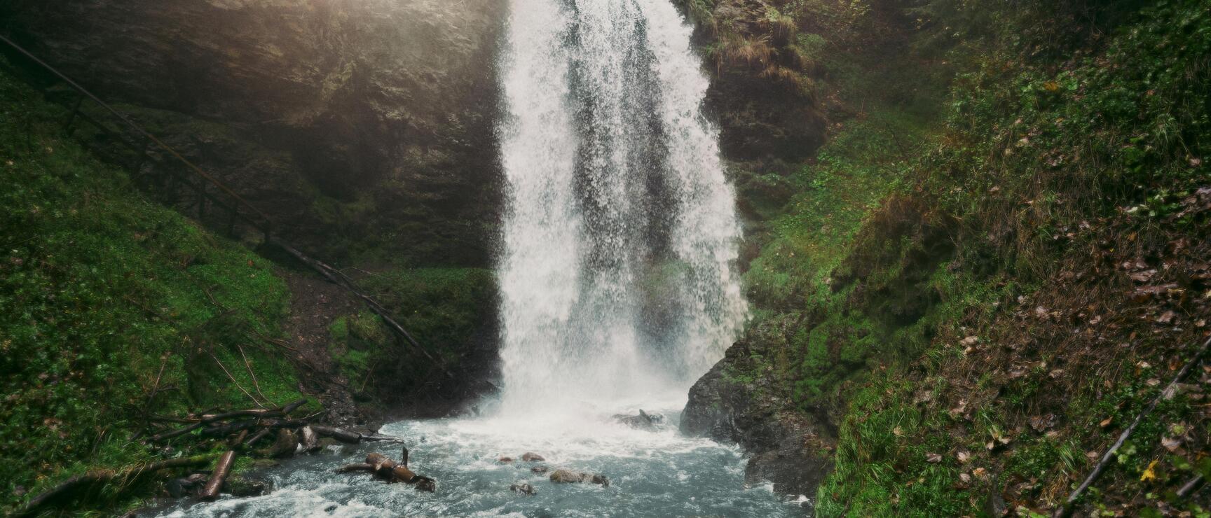 Tall waterfall cascading over rocks into a stream, surrounded by green forest and sunlight.