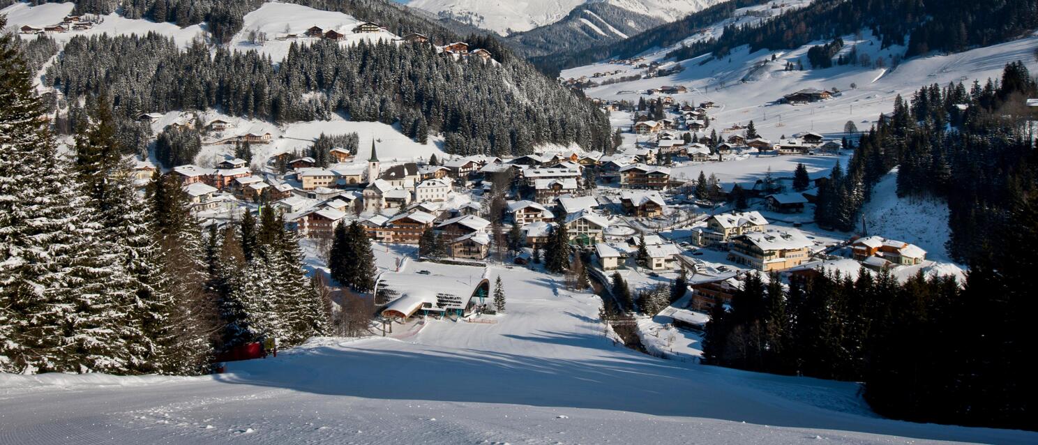 Wintry Alpine village with church tower in snowy valley, snow-capped mountain peaks and blue sky.