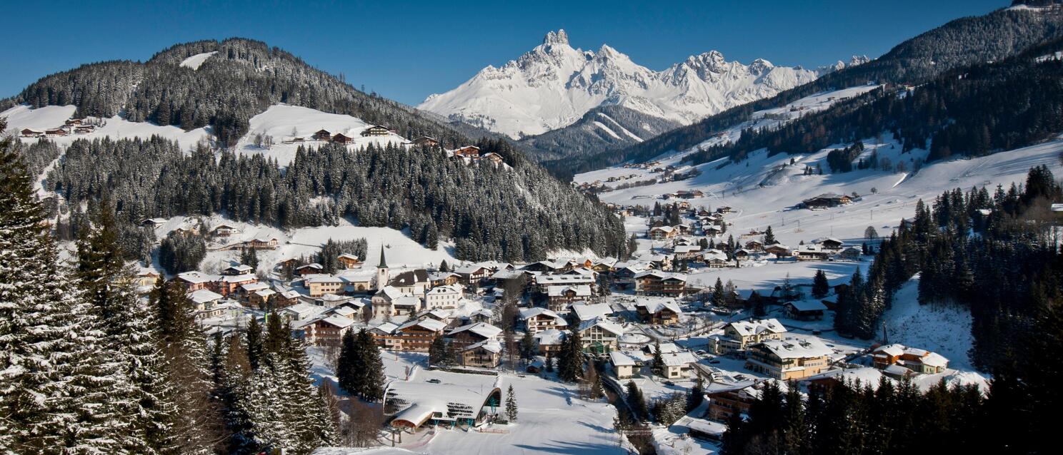 Winterliches Alpendorf mit Kirchturm im verschneiten Tal, schneebedeckte Berggipfel und blauer Himmel.