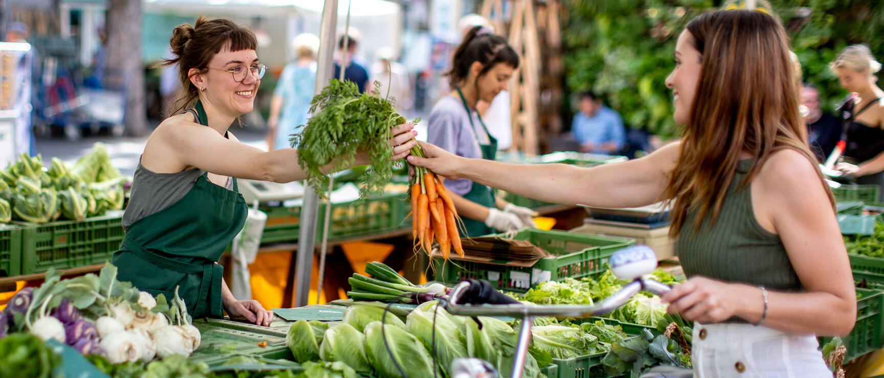 Market stall with vegetables: vendor in green apron handing bunch of carrots to customer, bicycle in foreground.