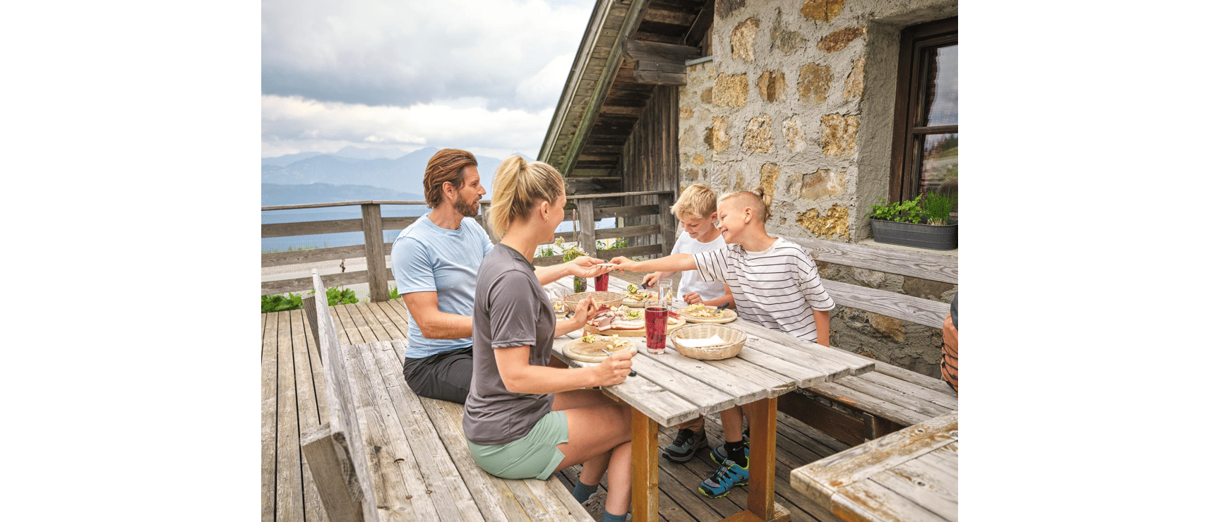 Zwei Erwachsene und zwei Kinder beim Essen auf der Terrasse einer Almhütte, Bergpanorama im Hintergrund.