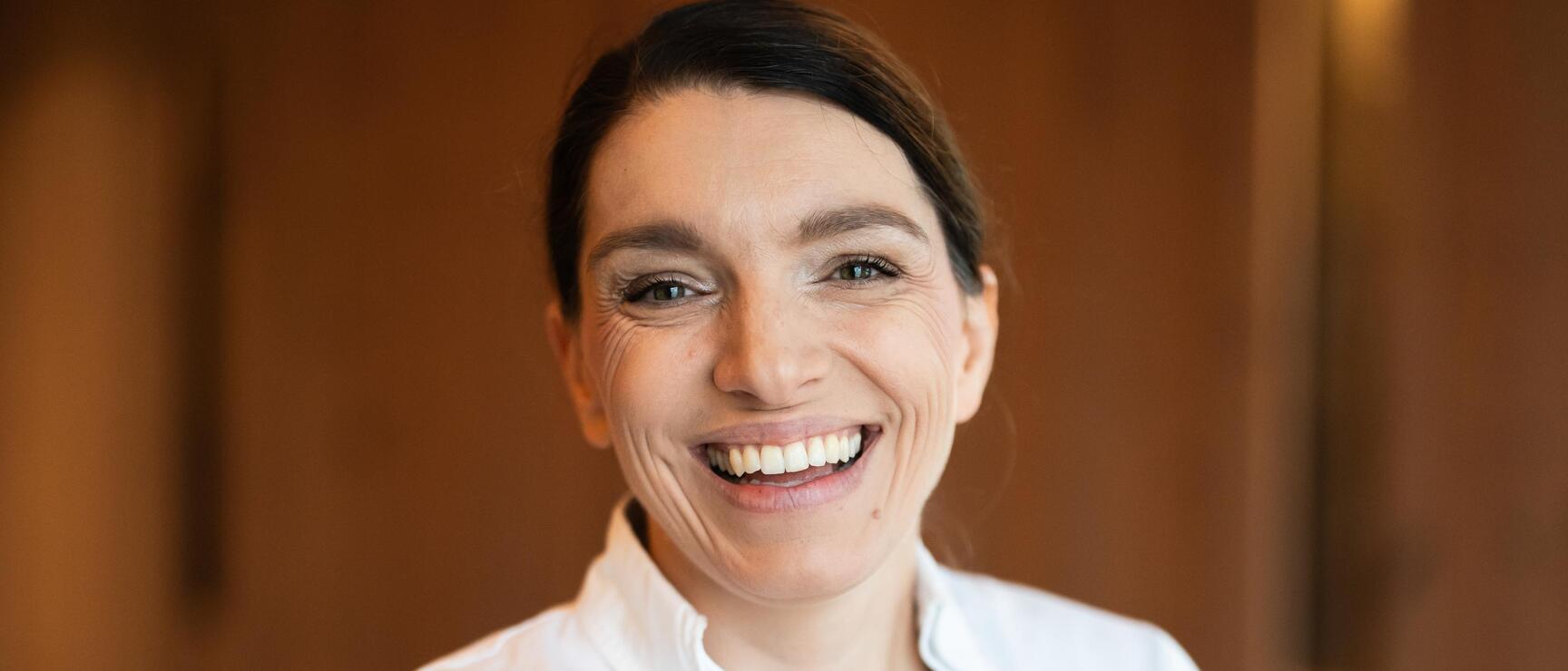 Smiling woman in white shirt with "Eveline Chocolatier" logo, wooden wall and lamp in background.