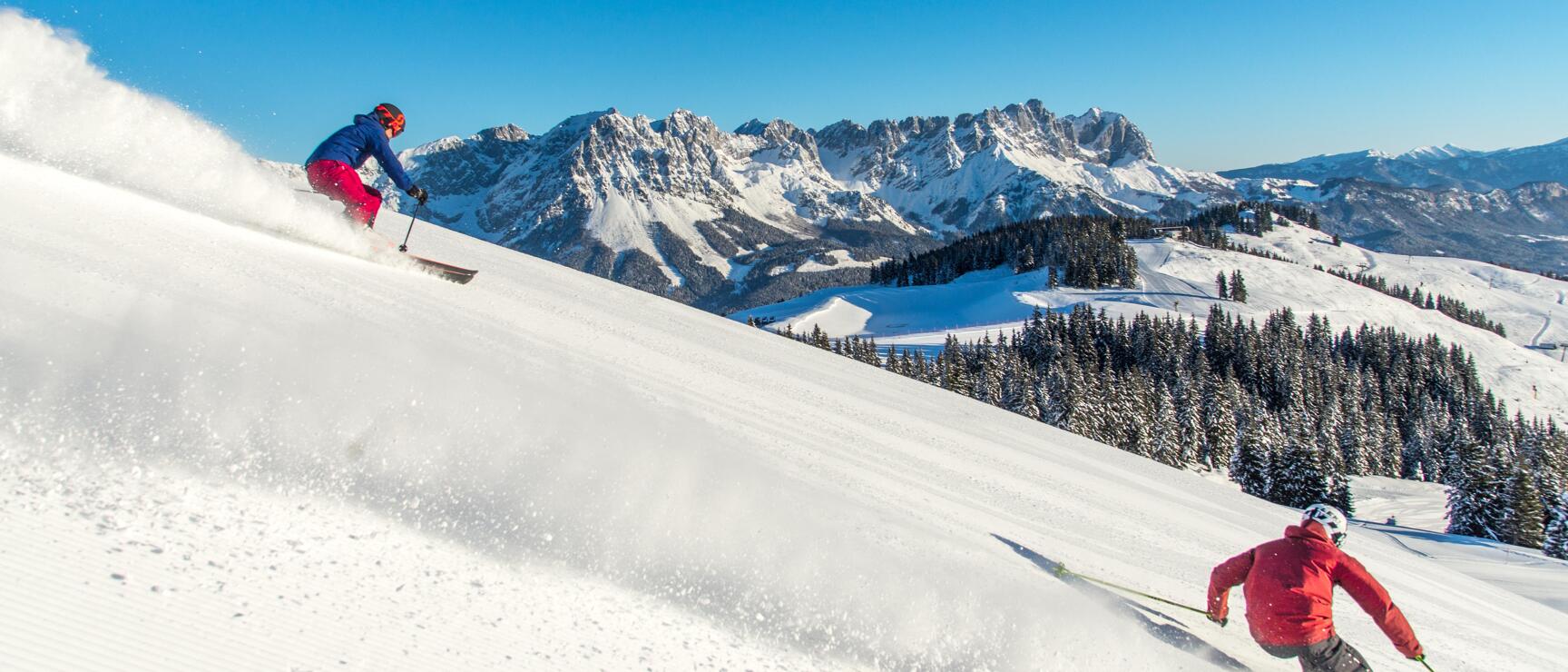 Two skiers on snowy slope, powder snow, snow-covered mountain ridge and coniferous forests in background