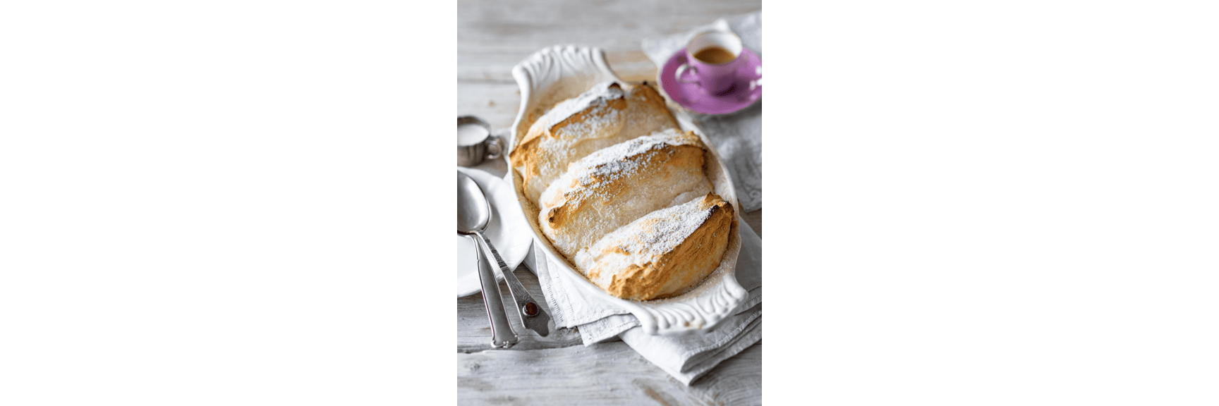 Salzburg nockerl with powdered sugar in white baking dish, beside it spoon and espresso cup.
