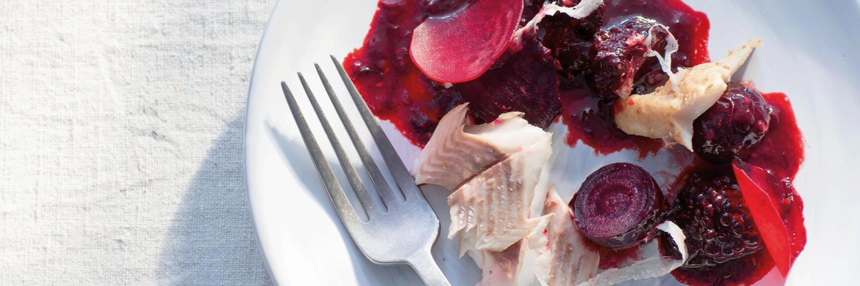 White plate with fish pieces, beetroot slices and berry sauce, fork beside, light linen cloth.