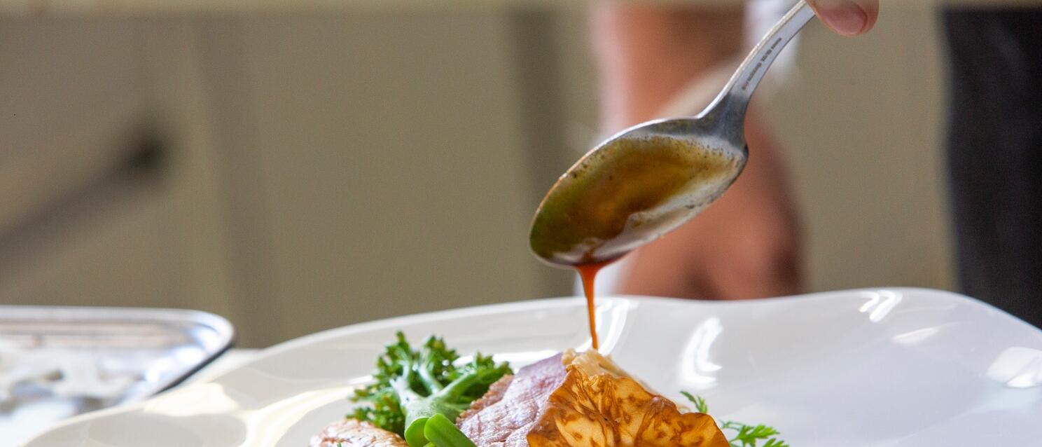 Close-up: meat dish with broccoli and dumplings on white plate, hand pouring sauce with spoon.