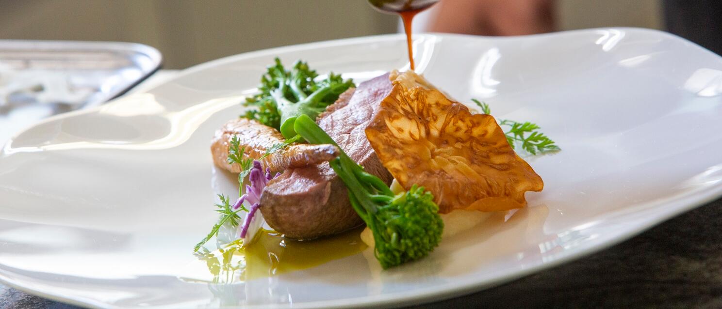 Close-up: meat dish with broccoli and dumplings on white plate, hand pouring sauce with spoon.