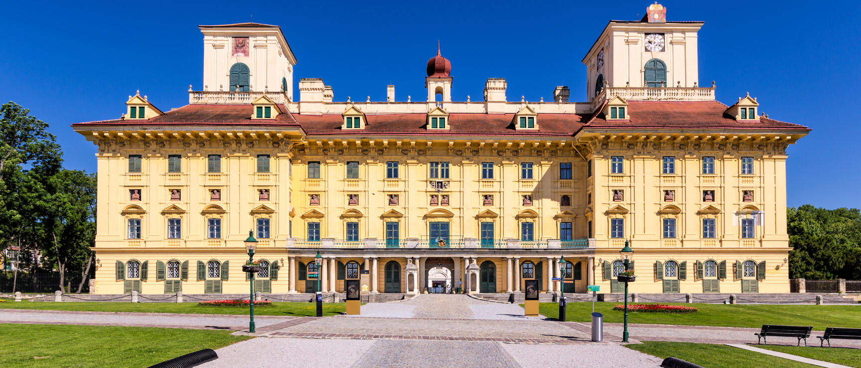 Esterházy Castle in Eisenstadt, yellow Baroque façade with red roof, gravel path and lawn in front, blue sky.