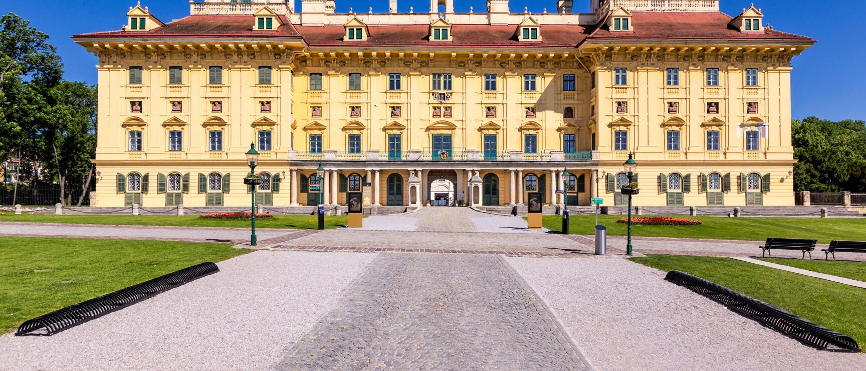 Schloss Esterházy in Eisenstadt, gelbe Barockfassade mit rotem Dach, Kiesweg und Rasenfläche davor, blauer Himmel.